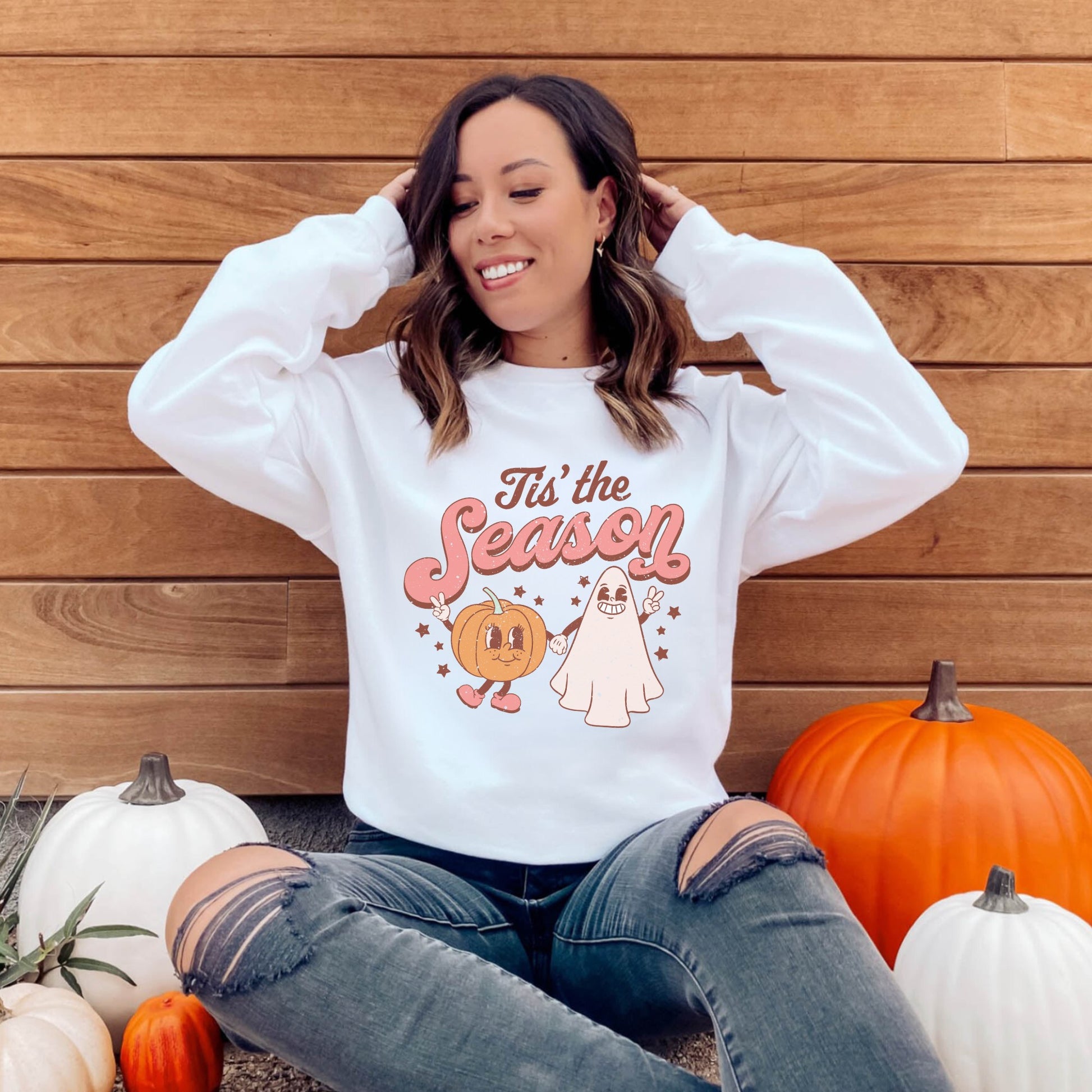 A retro, vintage Halloween sweatshirt crewneck with a cute ghost and pumpkin design. The sweatshirt features the slogan "Tis the Season" in bold, playful letters. The muted colors and vintage design add to the overall spooky aesthetic of the sweatshirt.