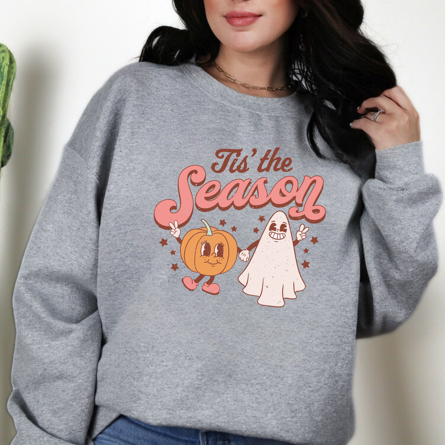 A retro, vintage Halloween sweatshirt crewneck with a cute ghost and pumpkin design. The sweatshirt features the slogan "Tis the Season" in bold, playful letters. The muted colors and vintage design add to the overall spooky aesthetic of the sweatshirt.